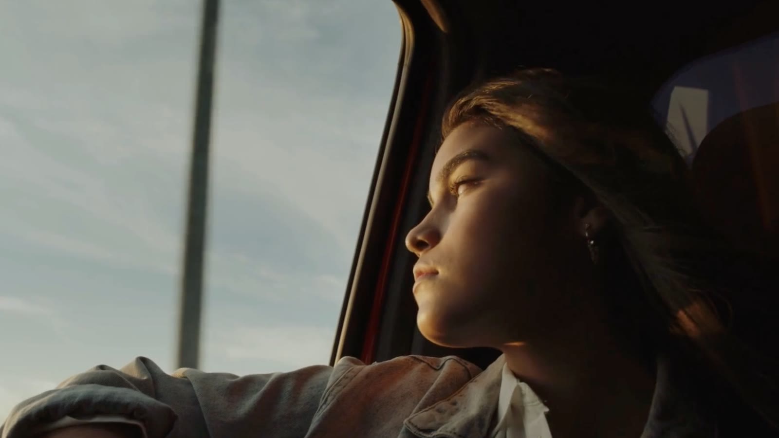 A young woman in a denim jacket looking out a car window at golden-hour sky